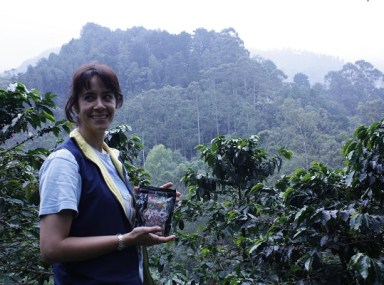 En kvinna står i en kaffeodling och ler. I handen har hon ett kaffepaket